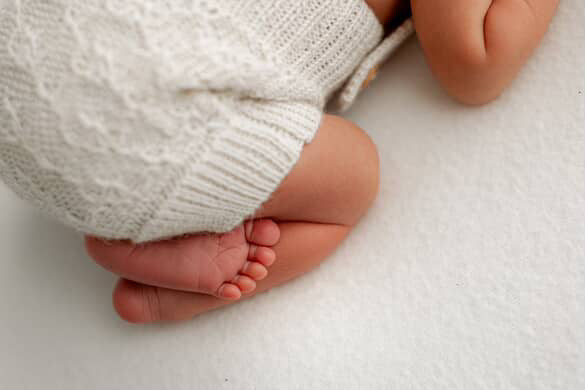 Close-up of a baby's legs and feet wearing white knitted pants on a white surface.