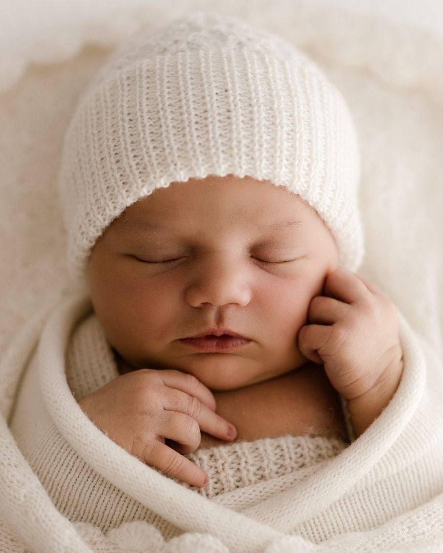 Newborn baby wrapped in white fabric and wearing a matching white knit hat.